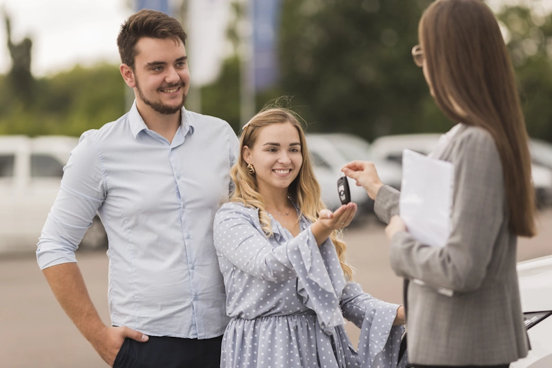 A man stands next to a young girl who is receiving car keys from a woman holding car title documents in a parking lot.