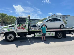 A man next to a tow truck with a white car loaded, advertising cash for junk cars in Margate, FL.