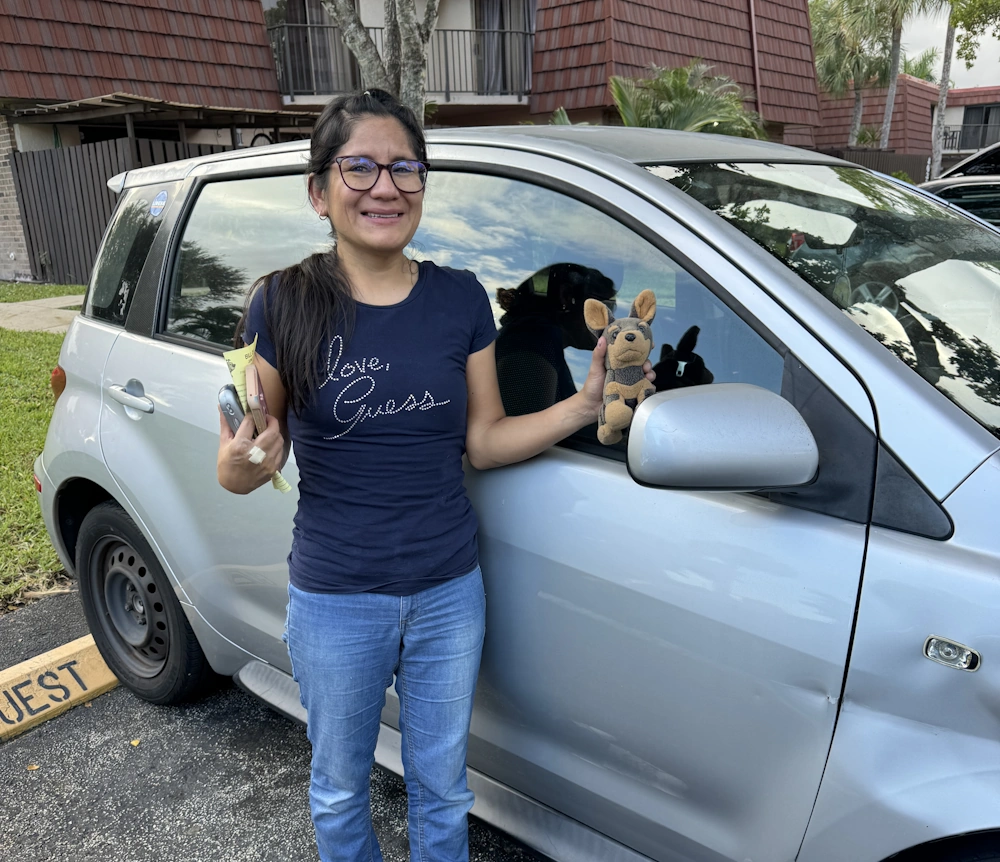 Woman standing beside a silver car holding a plush toy and keys representing vehicle buyers in Broward County, FL.
