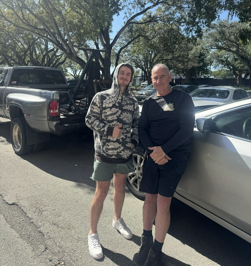 Two men standing next to a silver car and a tow truck for Junk Car Dog's we buy junk cars in Sunny Isles Beach service.