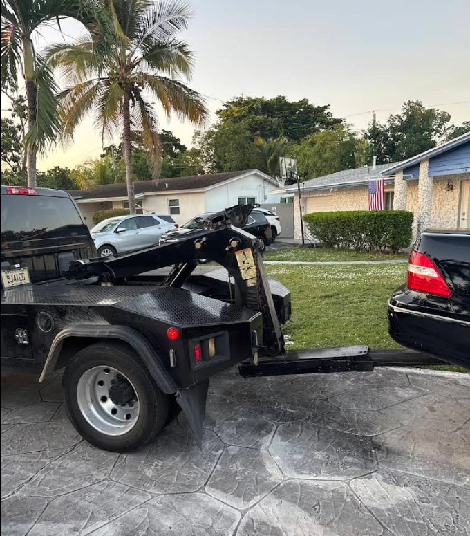 A black tow truck hitched to a black car in a residential driveway for Junk Car Dog's we buy junk cars in Sunrise service.