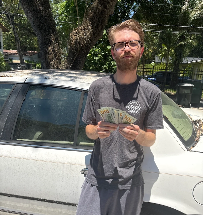 A man holding a fan of hundred-dollar bills while standing next to an older white sedan, showcasing cash returns from junk car options in Plantation FL.
