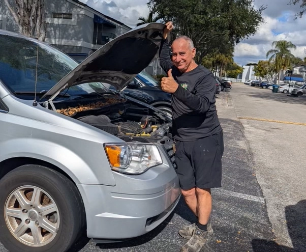 Man giving a thumbs-up beside a silver minivan with the hood open in a parking lot, representing a satisfied customer looking to sell my junk car in Fort Lauderdale.