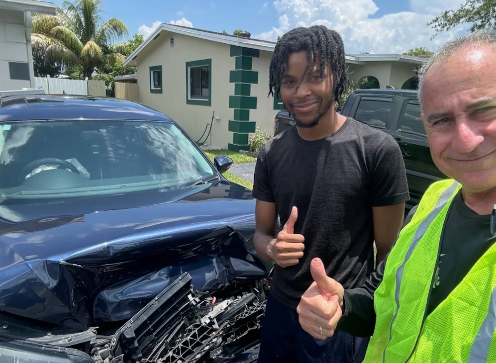 A young man and a Junk Car Dog representative stand beside a dark blue sedan with significant front-end damage, both providing a thumbs-up after finalizing the documents needed to junk a car.
